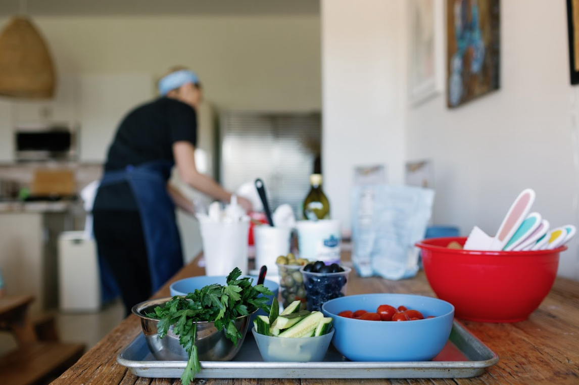 A table in our Olivenhain classroom with fresh vegetables like cherry tomatoes, cucumber slices, celery sticks, and herbs, with a person preparing food in the background.