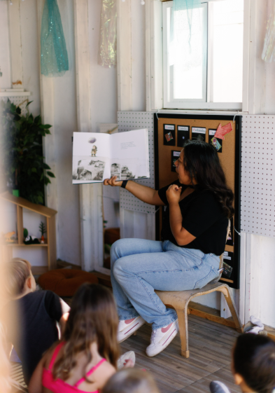 A preschool teacher reading a book to children in a cozy room with white walls, a window, and decorations.