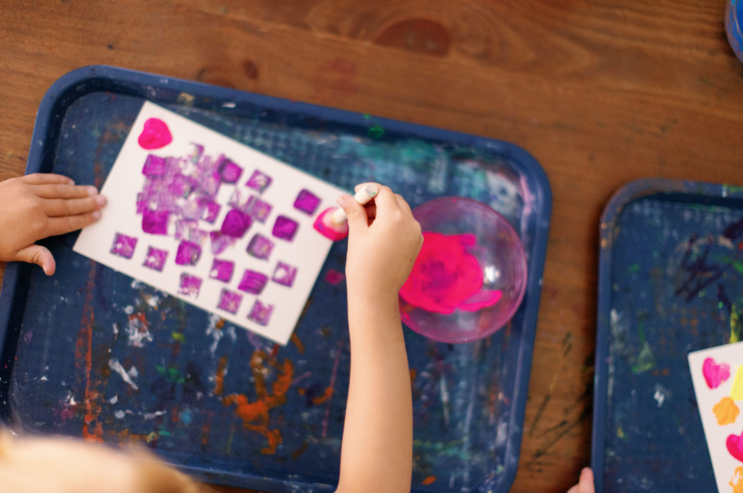 Child's hands creating artwork with bright pink and purple paint on a white paper.