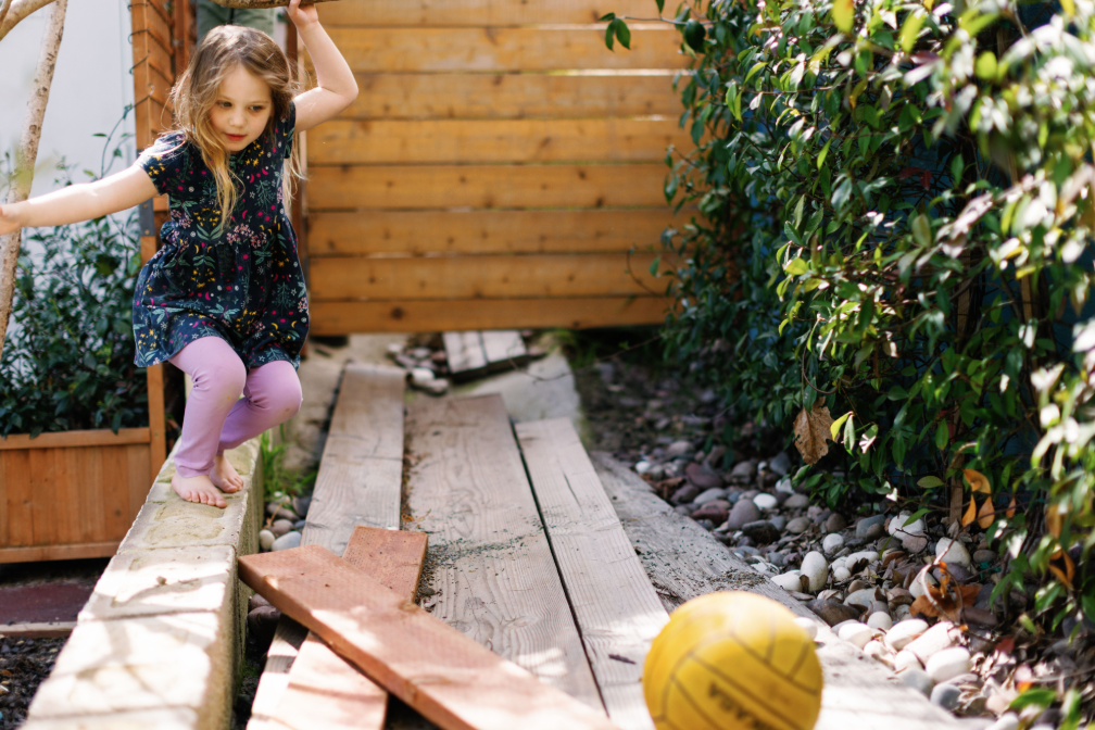 A young girl playing outside on a wooden plank path, wearing a floral dress and pink tights, with a yellow ball nearby in a preschool garden surrounded by greenery.