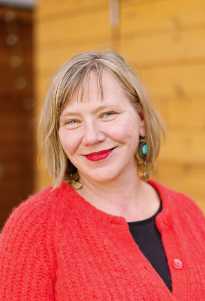 A woman with short blonde hair wearing a red cardigan and turquoise earrings smiling outdoors with a wooden background.