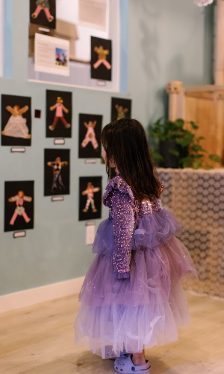 A young girl dressed in a purple, sparkly, layered tutu dress stands in front of a wall displaying various colorful art projects.