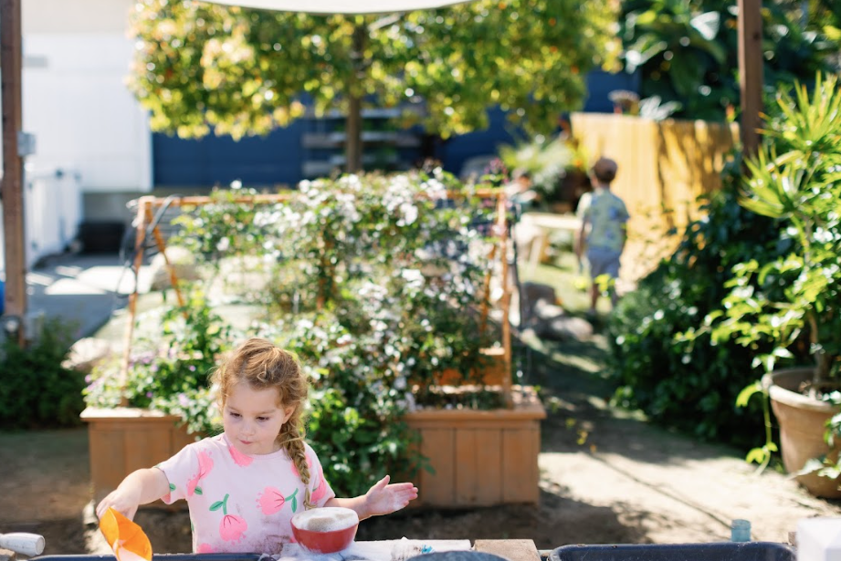 Young girl with curly hair wearing a pink shirt with floral pattern playing outdoors in a garden with greenery and flower beds
