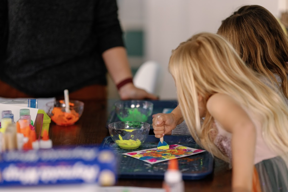 Two young girls engaged in finger painting at a table with art supplies, while an adult stands nearby.