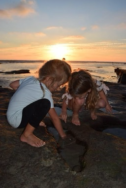Two young girls playing on rocks by the ocean at sunset, exploring a small tide pool.