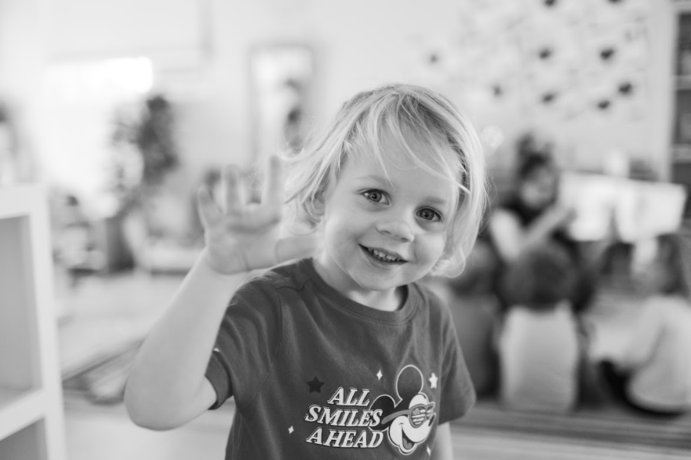 A young blonde boy smiling and making a peace sign with his right hand, in a bright indoor setting.