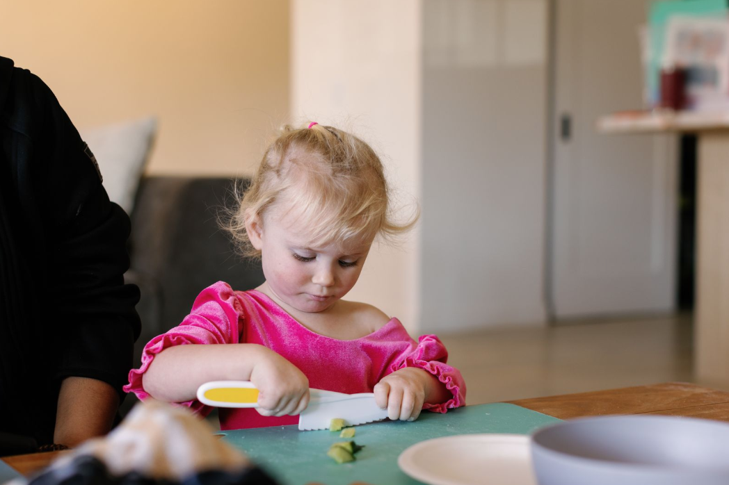 A young girl with blonde hair in a pink dress is cutting vegetables with a plastic knife at a kitchen table.