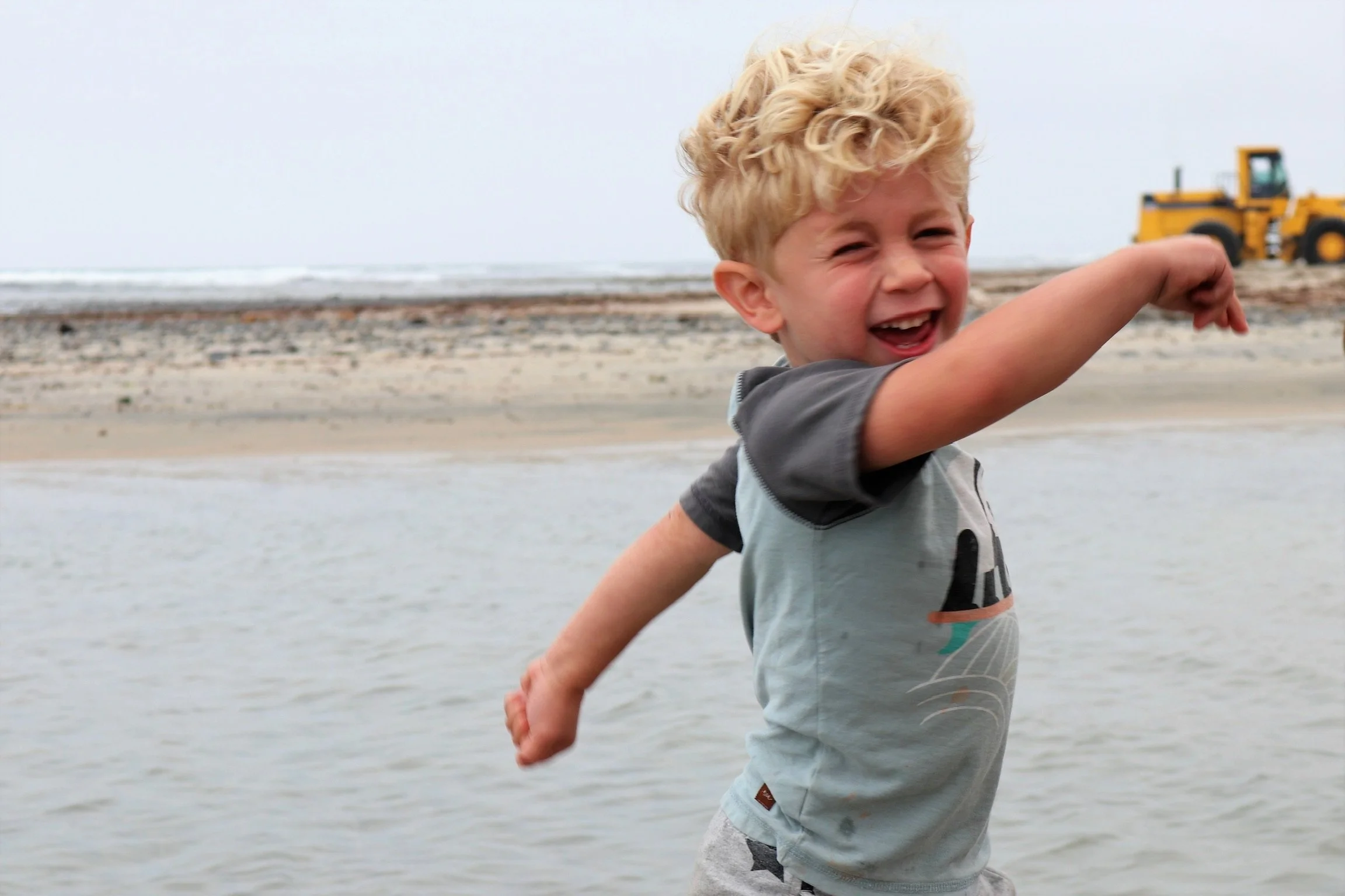 A young boy with curly blonde hair smiling and pointing at the ocean at the beach, with a yellow bulldozer in the background.