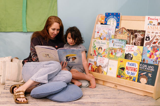 A teacher and a child sitting on the floor next to a bookshelf filled with children's books, reading a book together.