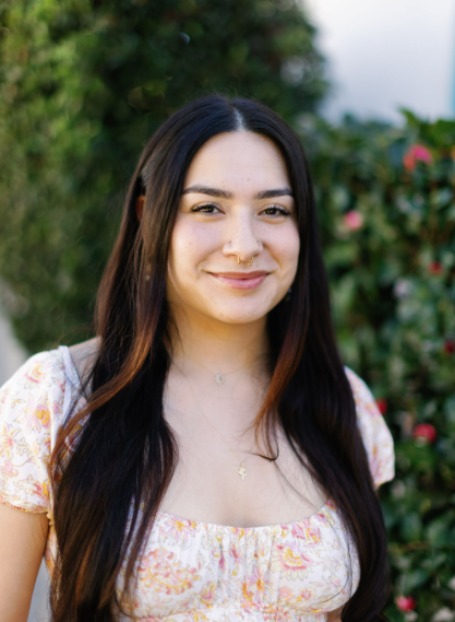 Young woman with long dark hair, smiling, wearing a floral top, standing outdoors with greenery and pink flowers in the background.