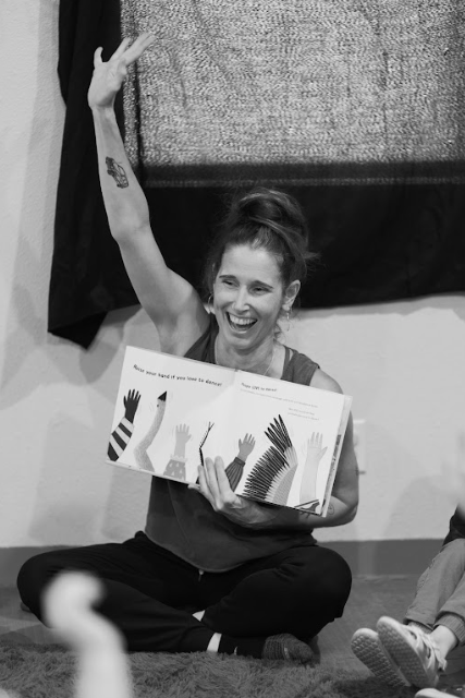 A woman sitting on the floor, smiling and raising her right hand, holding a children's book in her left hand.