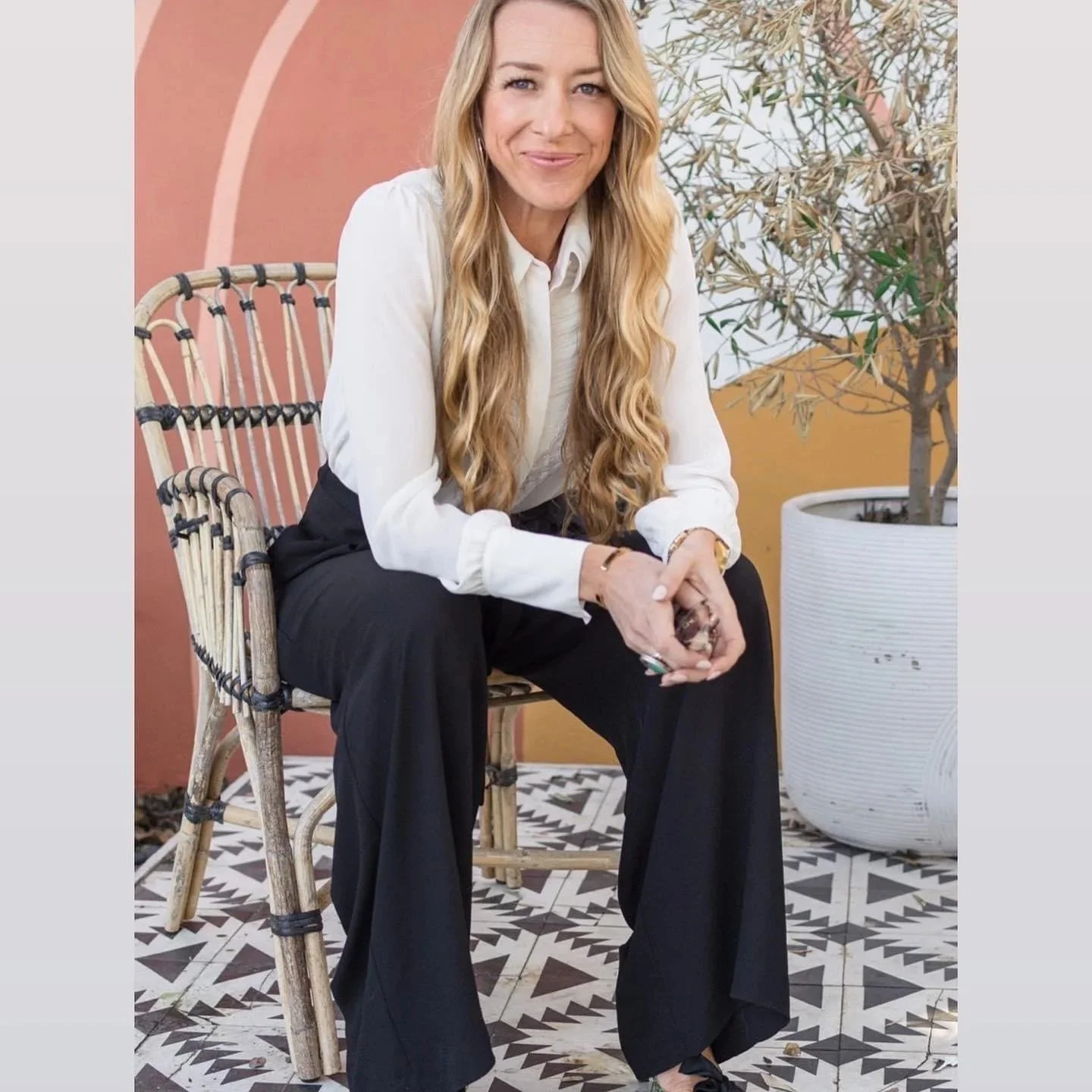 Casey Falkner, Kids By The Sea leadership, wearing a white blouse and black pants, sitting on a rattan chair with her hands clasped.