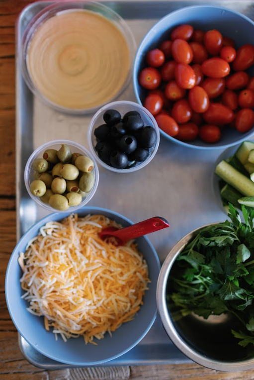 Bowls of cherry tomatoes, green olives, black olives, shredded cheese, and fresh herbs on a tray.