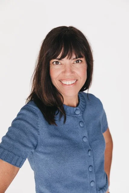 Kids By The Sea leadership, Erin Lane, a woman with dark brown hair and bangs smiling, wearing a blue short-sleeve button-up sweater, standing against a white background.