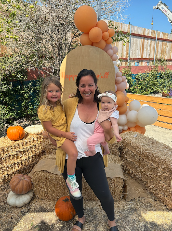 Woman holding two young girls in an outdoor setting with autumn decorations including pumpkins, hay bales, a wooden backdrop with balloons, and a tree with some green leaves, signifying a fall celebration.