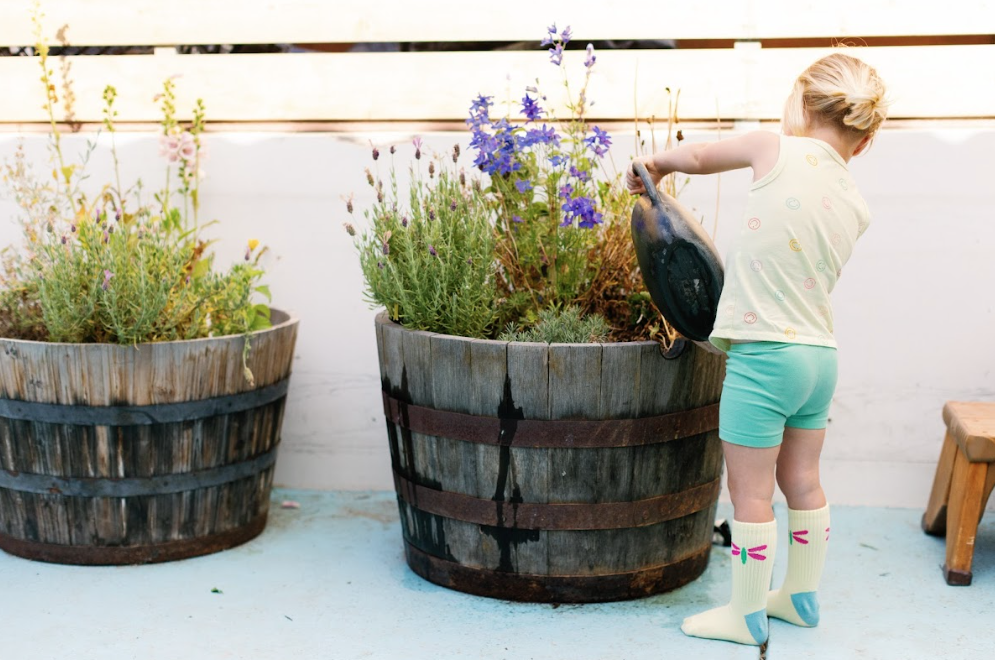 A young girl watering flowers in large wooden planters on a patio.