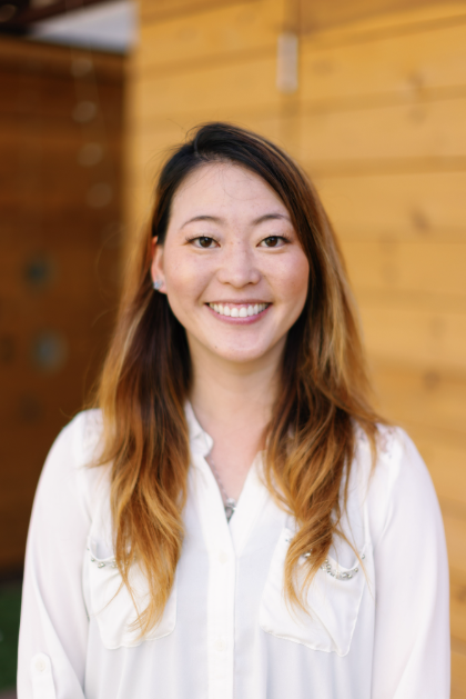 A smiling woman with long brown hair wearing a white blouse, standing in front of a wooden wall.
