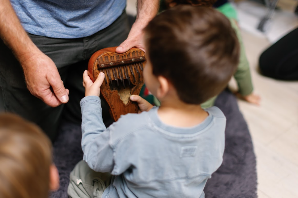 Young boy holding a wooden pan flute while an adult, Mr Troy, demonstrates how to play it, surrounded by other children on a floor.