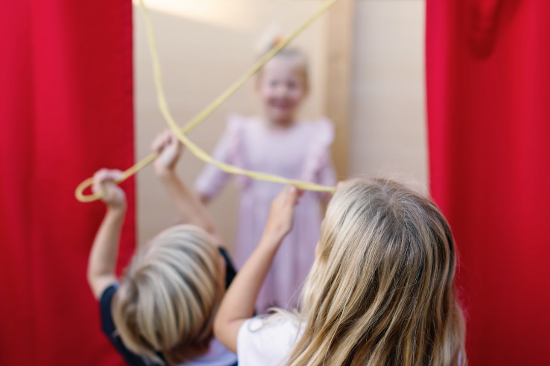 Two children use a yellow jump rope in front of a red curtain, while a girl in a lavender dress poses in the background.