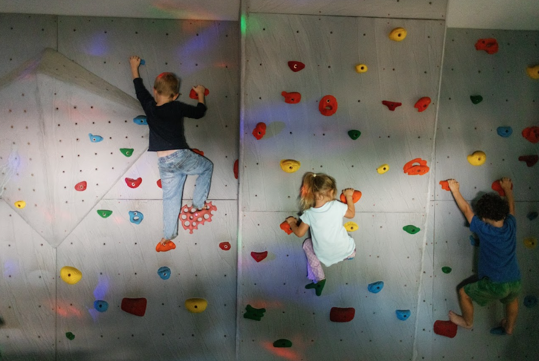 Three children climbing an indoor rock wall with multicolored holds.