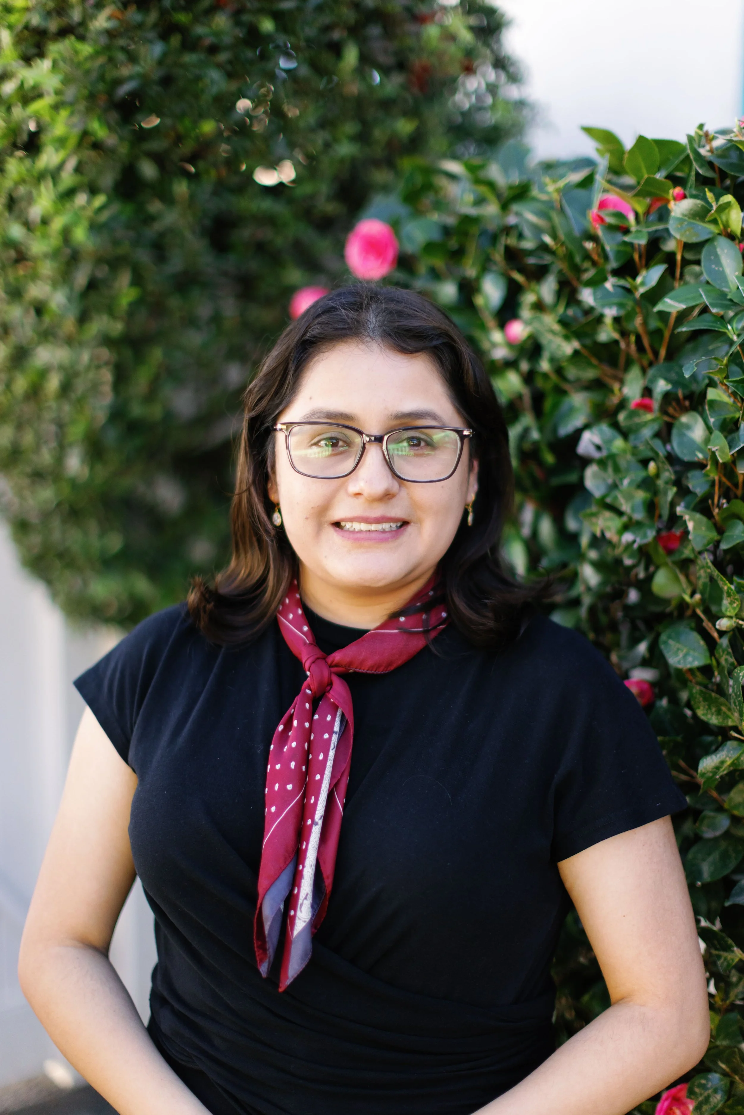 A woman with glasses and dark hair smiling outdoors, wearing a black shirt and a red polka dot scarf, standing in front of a bush with pink flowers.