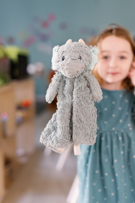 Young girl in a teal polka dot dress holding a gray plush dragon toy in a preschool playroom.