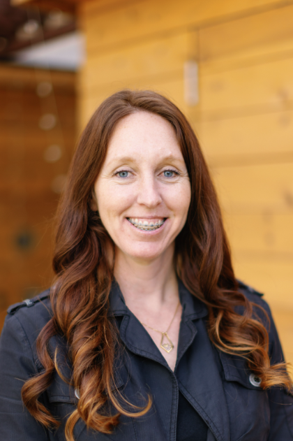 A smiling woman with long wavy red hair, wearing a black jacket, standing in front of a wooden wall.