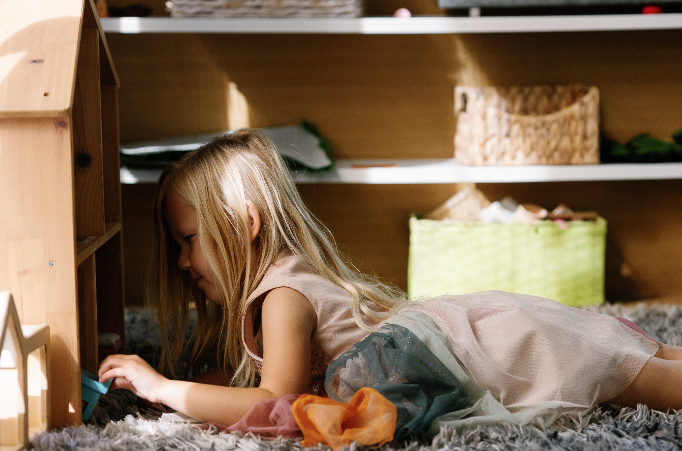 A young girl with blonde hair is lying on her stomach on a shaggy rug, playing with toys inside a wooden playhouse.
