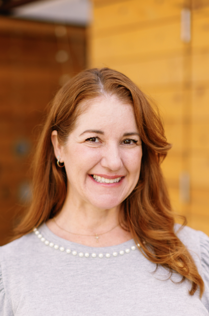 Woman with red hair smiling, wearing a gray top with pearl neckline, standing in front of wooden background.