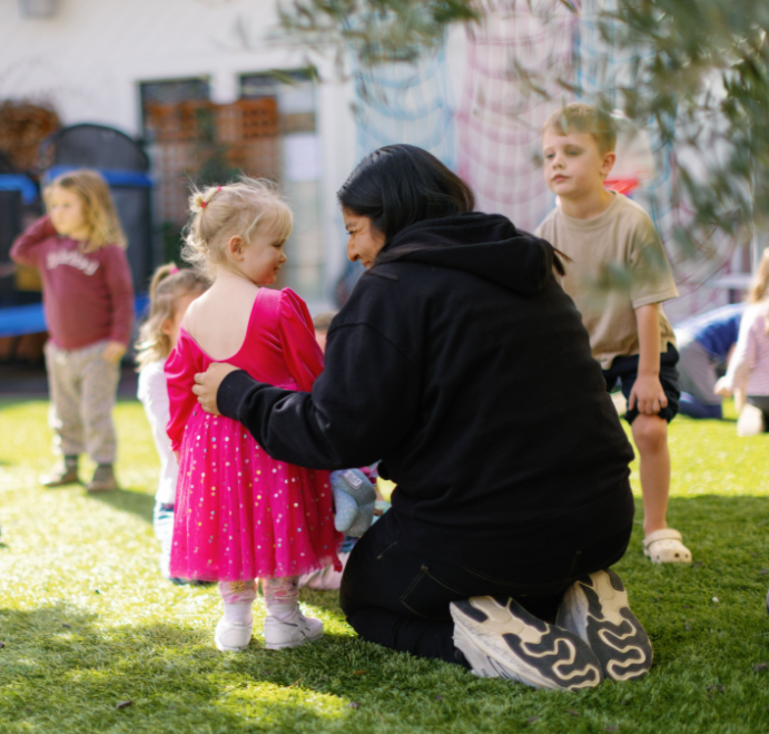 Preschool teacher kneeling on grass, interacting with a young girl in a pink dress, children playing outdoors on a sunny day.