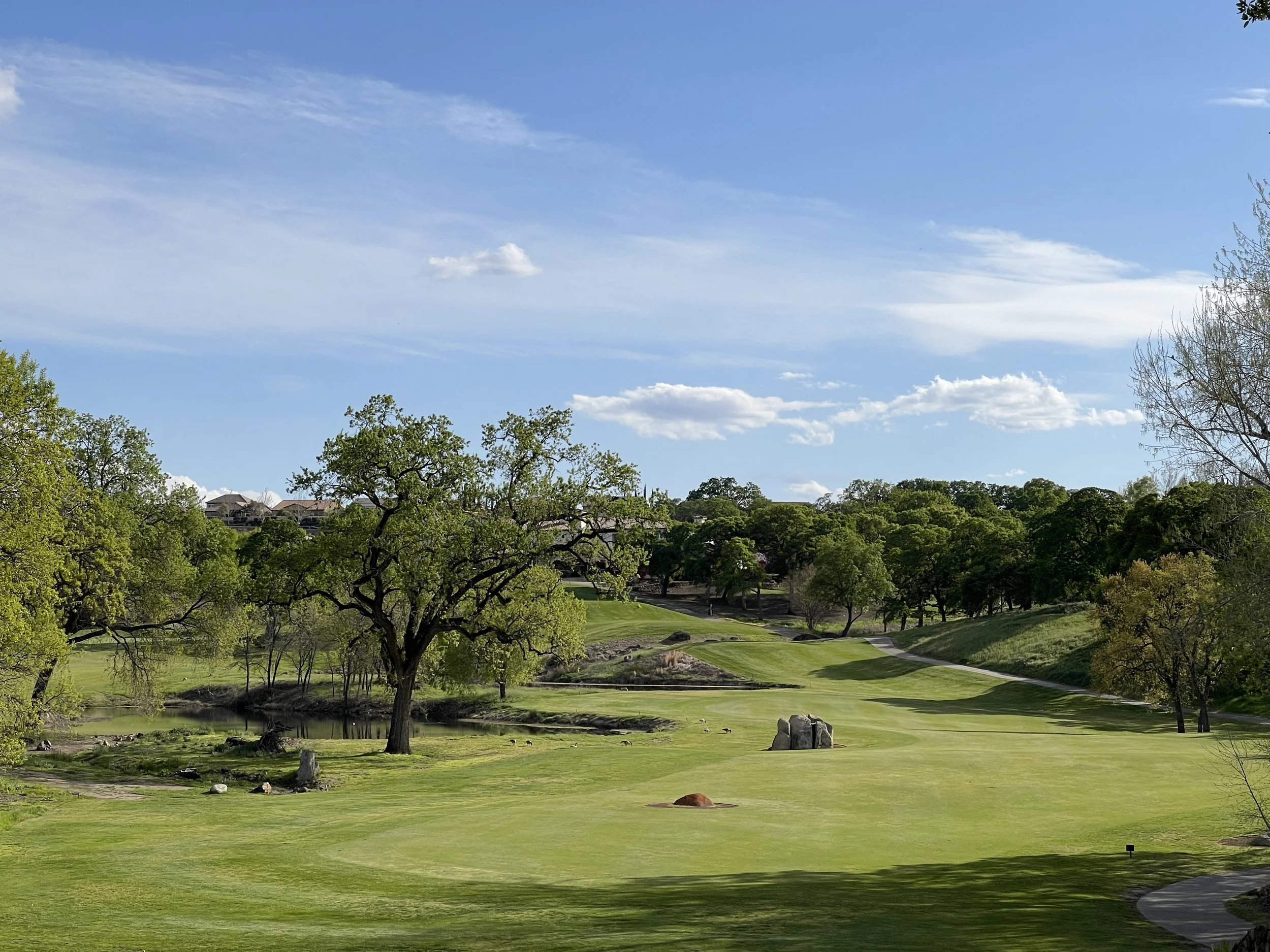 A scenic view of a golf course with green fairways, large trees, rocks, and a small pond. The sky is mostly clear with some clouds.