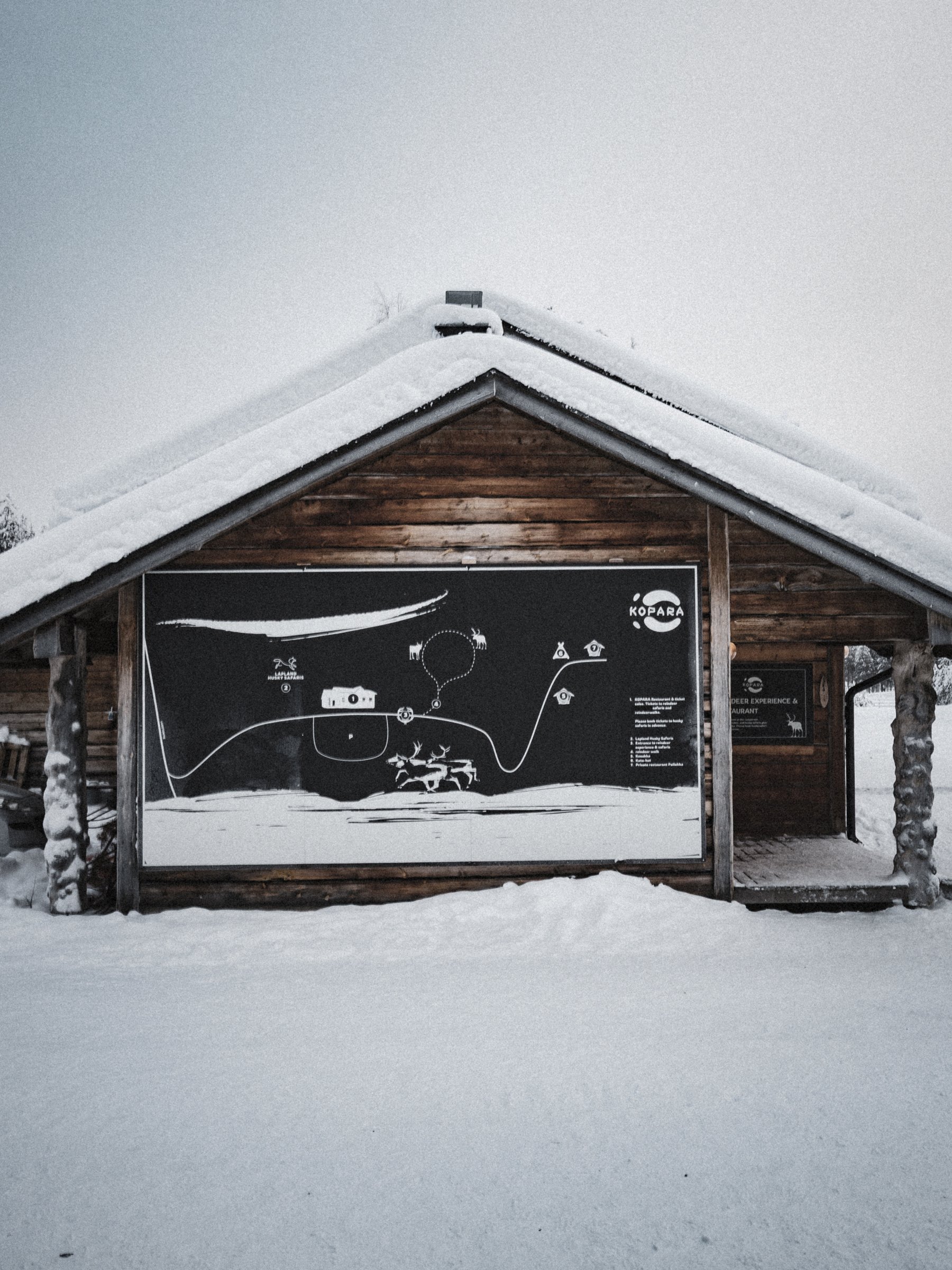 Ladina puinen maja lumisessa maisemassa, juliste seinällä, joka kuvaa talvisia aktiviteetteja ja alueen karttaa.