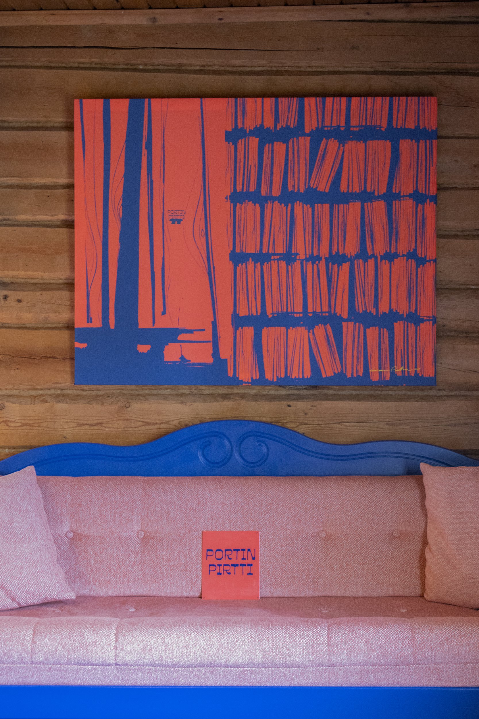 Kuva, joka sisältää vaaleanpunaisen sohvan ja sinisen puisen selkänojan välikön, taustalla lamellipaneelit ja sisätiloihin kuuluva maalattu taulu, jossa on kirjaimia ja kuvioita.