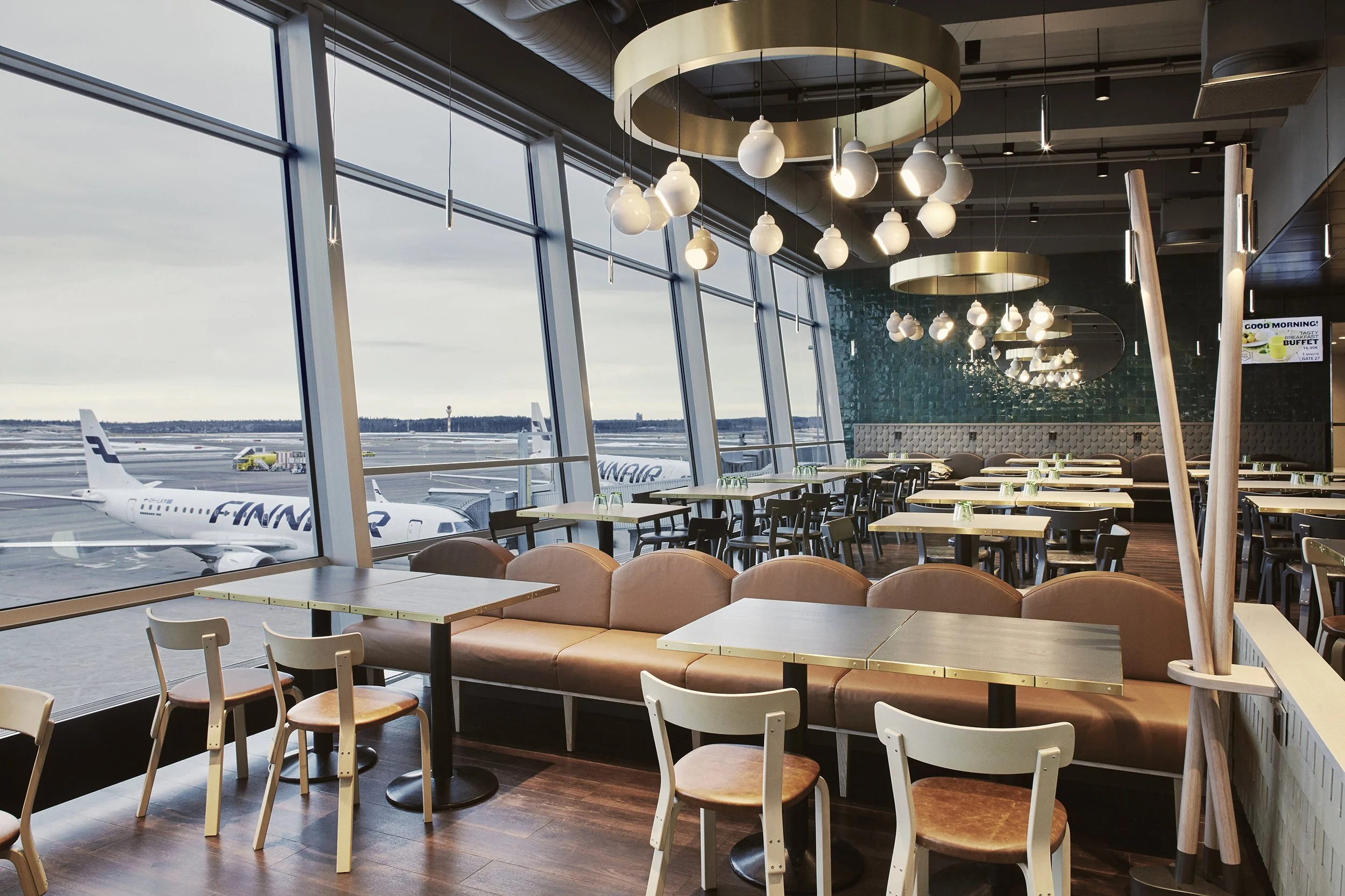 Lentoasema odotustilassa ikkunoista näkymä koneisiin ja lentokentälle, moderni sisustus, pöytiä ja tuoleja, kattovalaisimia.