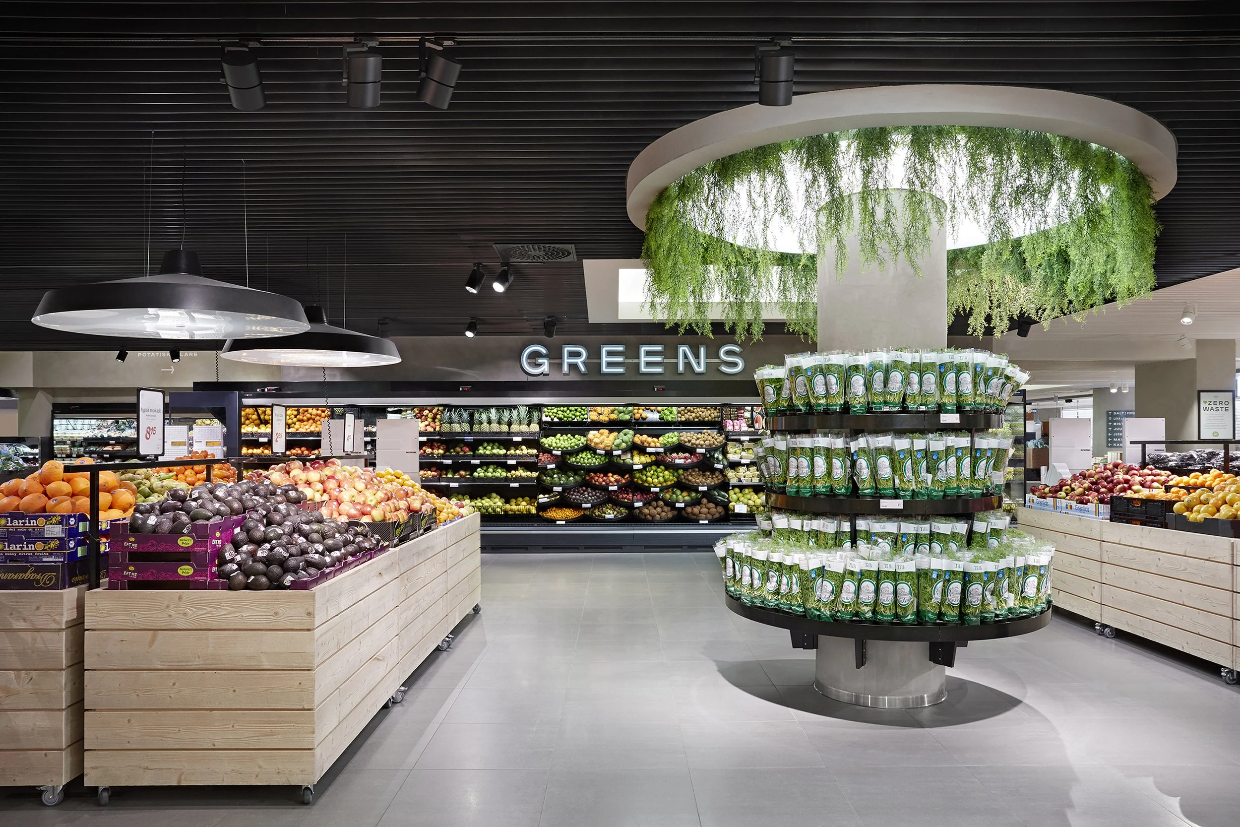 Vihannestorit ja hedelmät osastolla ruokakaupassa, otsikko 'GREENS' taustalla, vihreitä kasveja katossa, myymälä nykyaikainen ja siisti.
