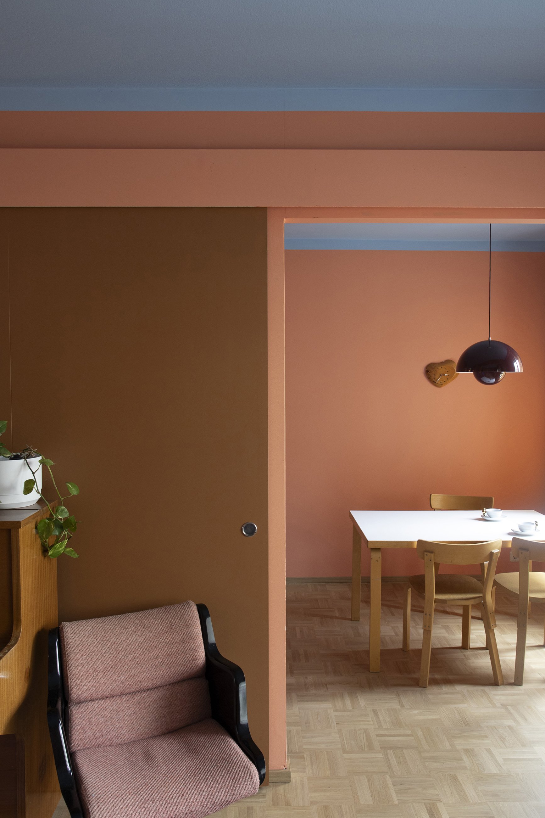 Kuvan sisätiloista, jossa on keskeneräinen ruokailutila ja nojatuoli vasemmalla. Pöytä ja tuolit ovat luonnonpuuta, seinät vaaleanpunaiset, ja kattoplafondi on silmiinpistävän tummanruskea.