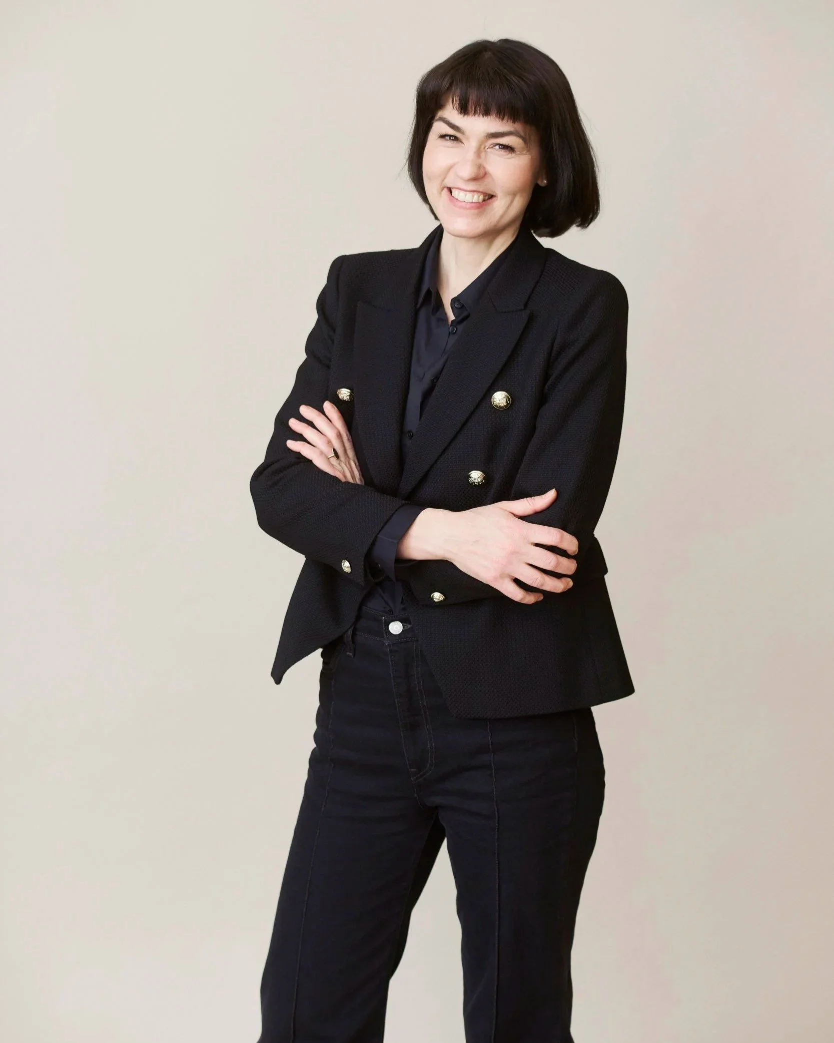 Nainen hymyilee ja seisoo rinta rinnan, hänellä on musta puku ja harmaat housut, taustana vaalea seinä.