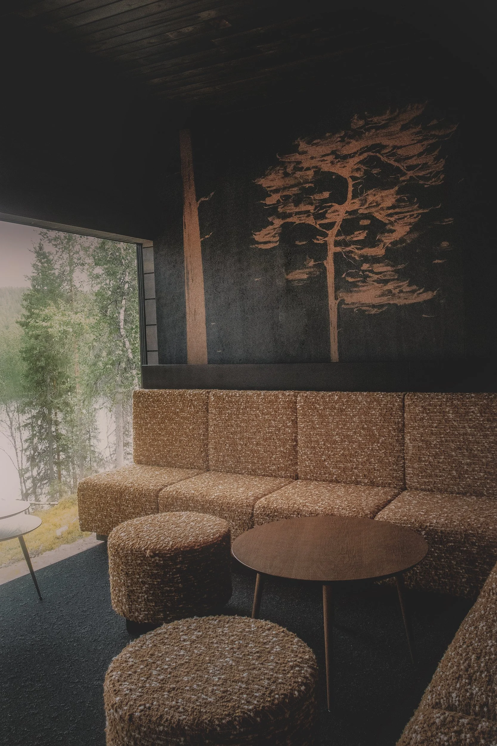 Sisätiloissa, suurilla ikkunoilla, käsintehtyä kankaalla verhoiltu sohvaryhmä ja pöytiä, vakava puutaideteos seinällä, luonto näkyy ulkona.