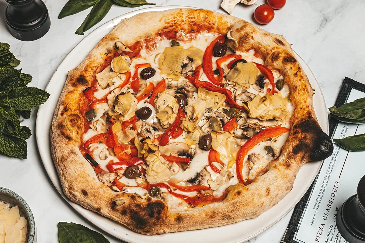 Une pizza avec garnitures de jambon, champignons, olives noires, poivrons rouges et artichauts, sur une assiette blanche.