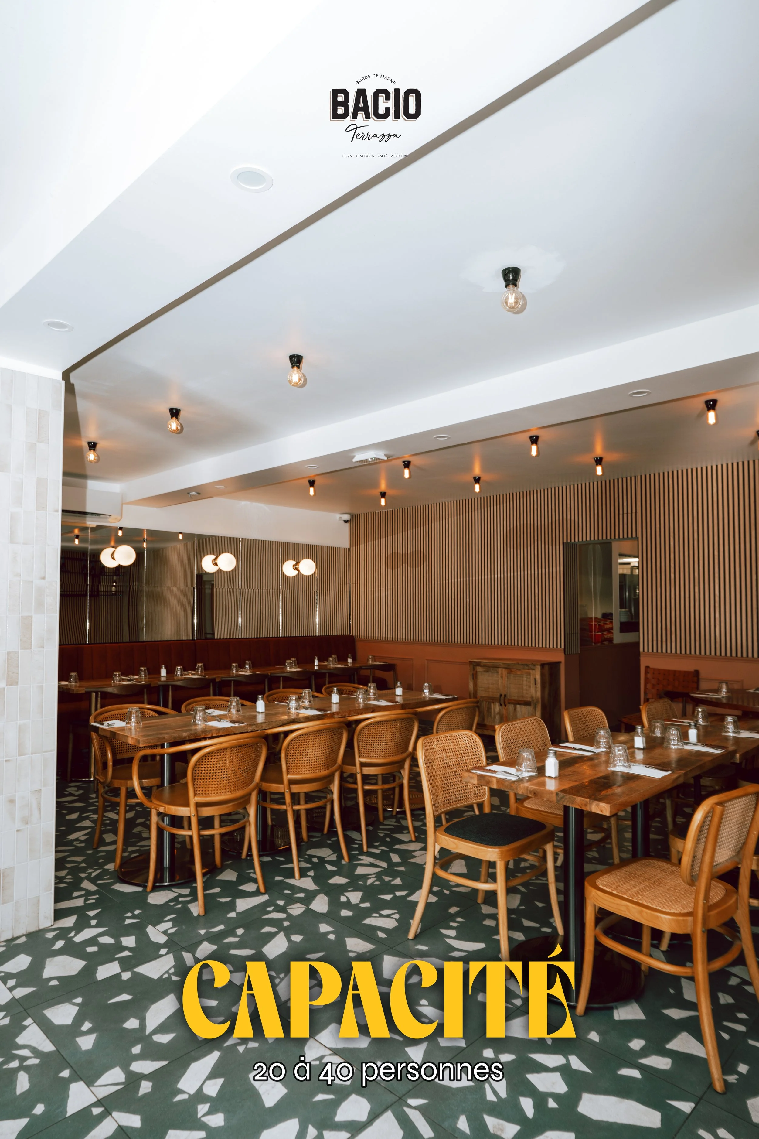 Intérieur d'un restaurant avec tables en bois, chaises en rotin, décor moderne, éclairage suspendu, sol à motifs géométriques, panneau sur le mur avec le nom 'Bacio' et l'inscription 'Capacité 20 à 40 personnes'.