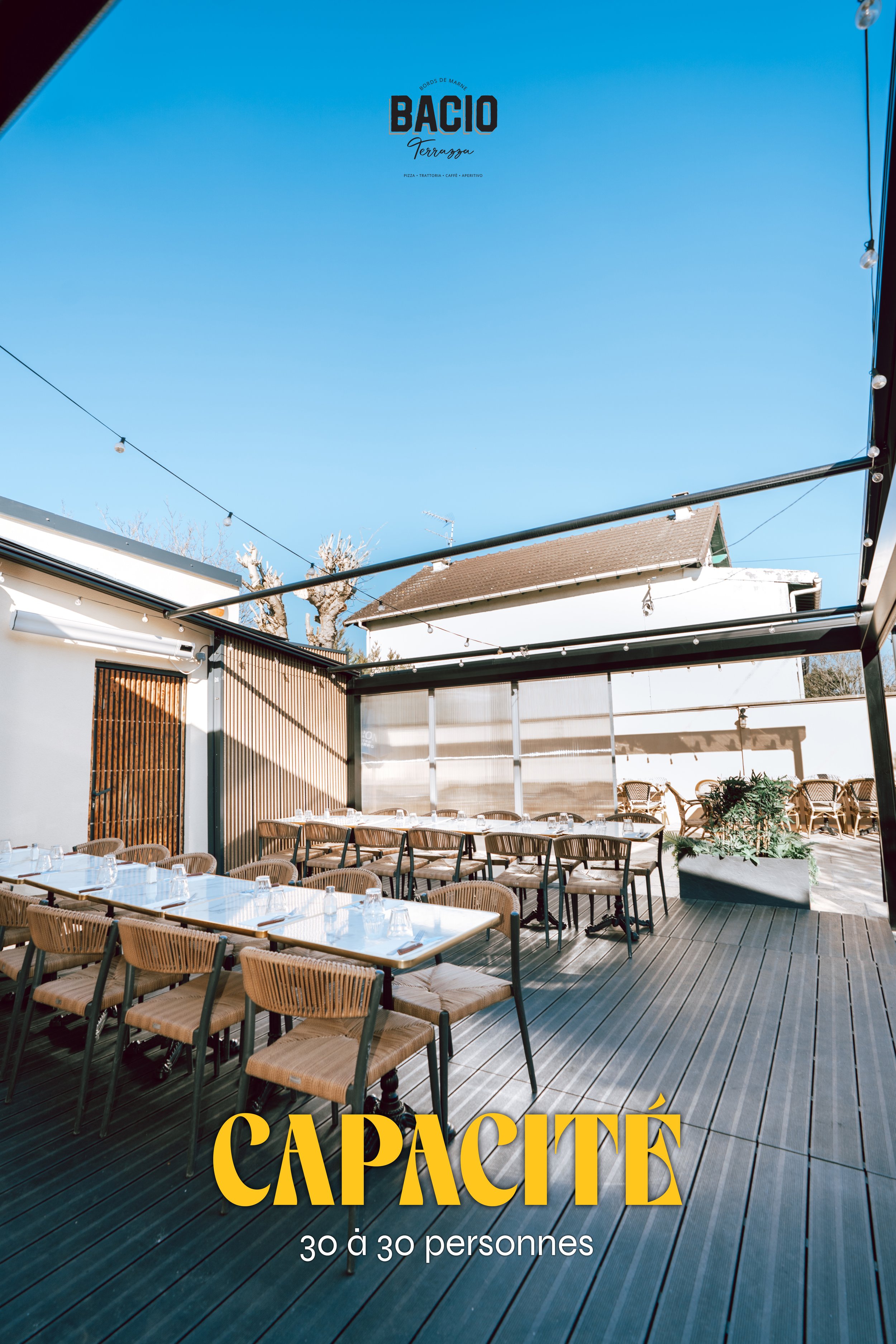 Une terrasse de restaurant en plein air avec plusieurs tables et chaises en rotin, des plantes vertes en pot, des éclairages suspendus, un ciel bleu clair et une maison en arrière-plan. Texte sur l'image : 'CAPACITÉ 30 à 30 personnes'.