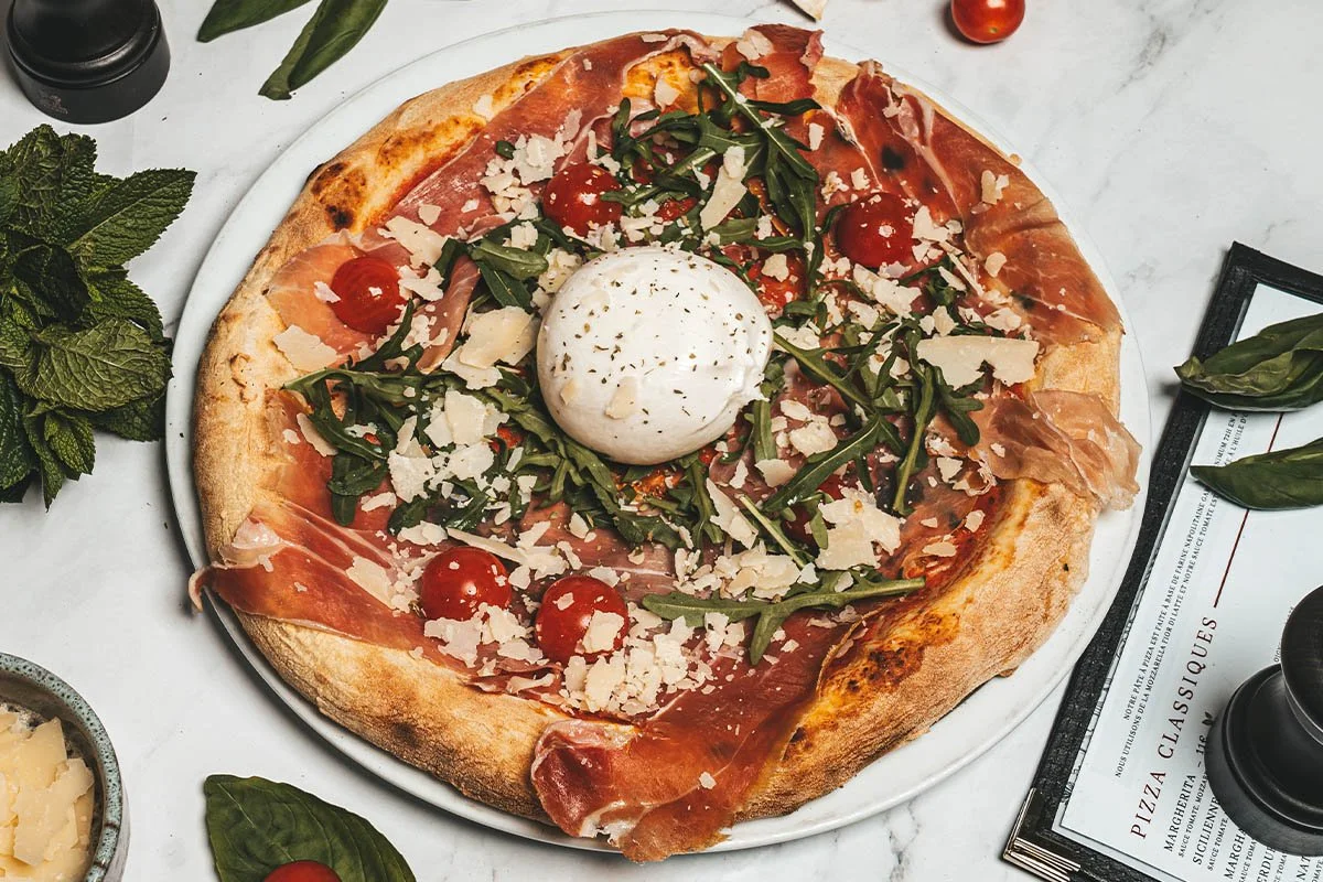 Pizza garnie de jambon, roquette, tomates cerises, copeaux de parmesan, et un œuf au centre, servie sur une assiette blanche.