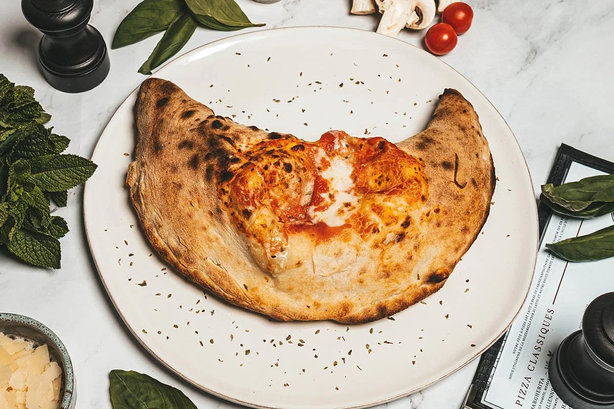 Une pizza calzone avec une garniture de fromage, tomate et œuf, servie sur une assiette blanche, entourée de feuilles de basilic, de menthe, de tomates cerises, de parmesan en morceaux et d'ustensiles de cuisine sur une surface en marbre.