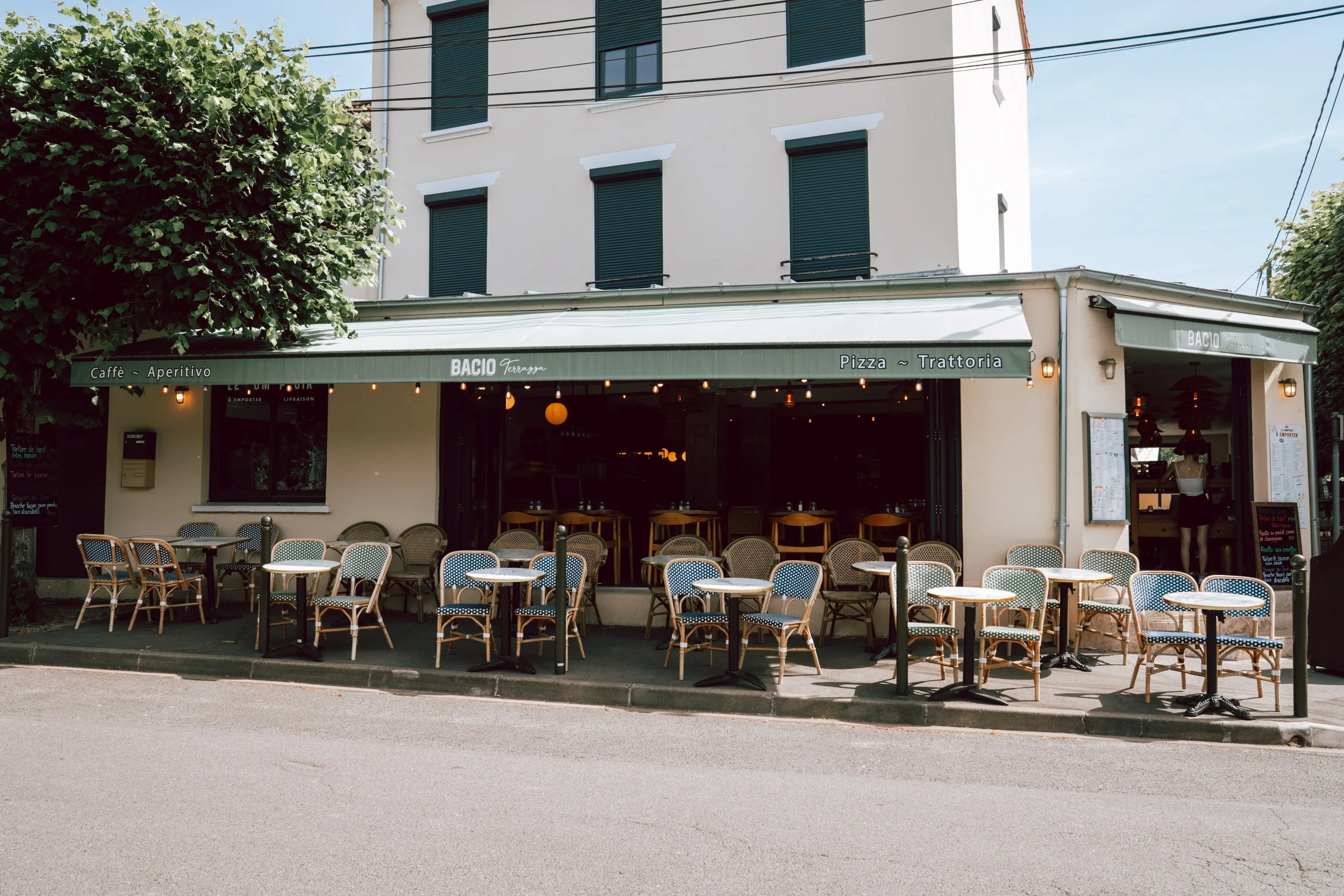 Une façade de restaurant avec une terrasse extérieure, des tables et des chaises en napperons, et un bâtiment résidentiel en arrière-plan. La terrasse porte une bannière indiquant 'Caffè – Aperitivo', 'Pizza' et 'Trattoria'.
