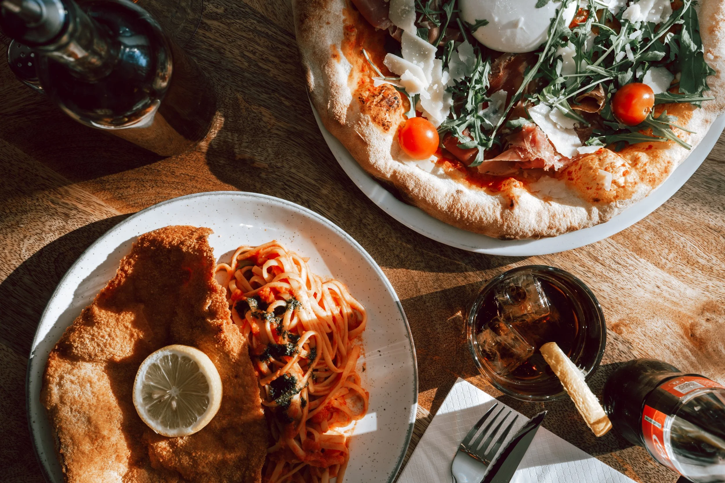 Une pizza avec des tomates cerises, du jambon, du fromage et des feuilles de roquette, une assiette avec un filet de poisson pané, des pâtes à la sauce tomate et un demi-citron, deux verres de boisson gazeuse avec des glaçons et une tranche de citron, un couteau, une fourchette, une serviette en papier et une bouteille de boisson gazeuse sur une table en bois