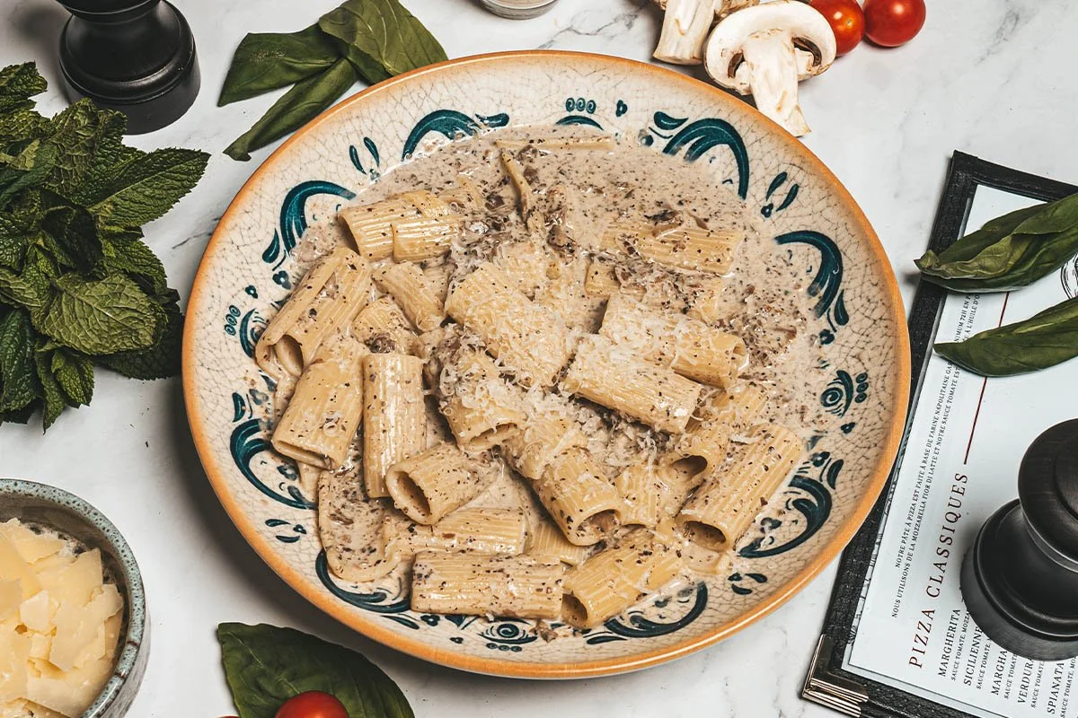 Plate de pâtes rigatoni avec une sauce crémeuse, probablement avec champignons et épices. Autour, il y a des feuilles de basilic, de la menthe, des tomates cerises, des champignons, du fromage râpé et des condiments.