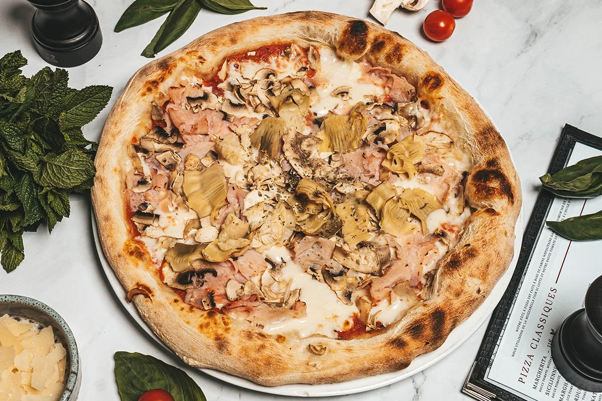 Une pizza aux champignons, jambon et fromage sur une table avec des tomates cerises, des feuilles de basilic et de la menthe