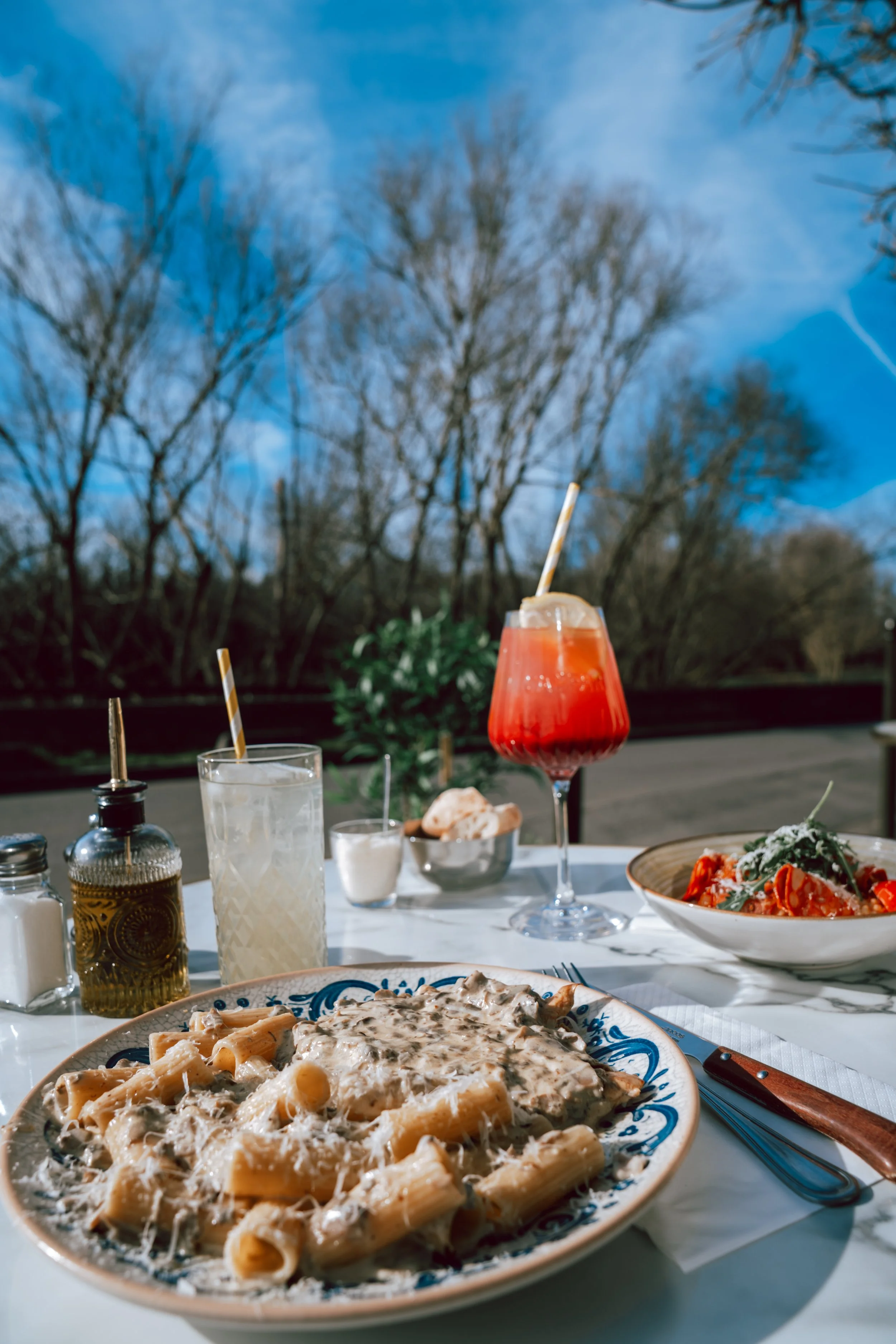 Un plat de pâtes à la crème avec des champignons dans une assiette bleue, une pizza à la sauce tomate avec du basilic, une boisson rouge avec une paille dans un verre à vin, et d'autres boissons et condiments sur une table, avec un ciel bleu et des a