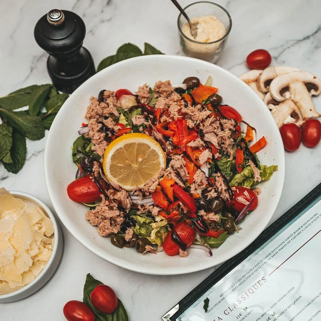 Salade méditerranéenne avec tomates cerises, thon, olives, et poisson, garnie d'une tranche de citron, avec du fromage râpé et des champignons en arrière-plan.