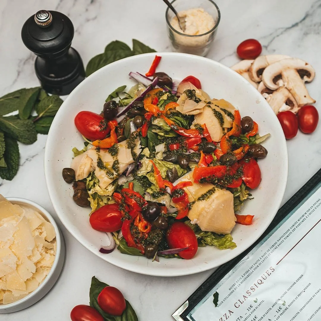 Salade composée avec tomates cerises, olives, morceaux de poulet, poivrons rouges, oignons, morceaux de champignons, accompagnée de parmesan et de sauce pesto.